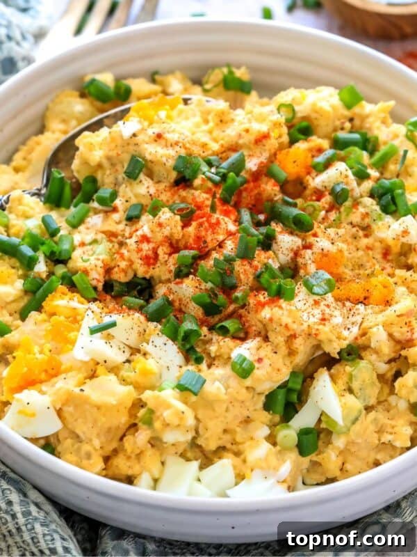 White ceramic bowl with deviled egg potato salad. Green onions on top and to the side with a spoon. Up close photo.
